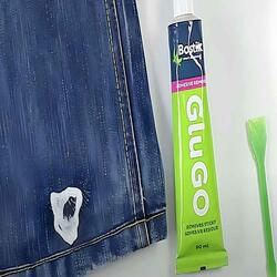A tube of Bostik Glu GO and a green scraper placed next to a denim jeans leg with chewing gum applied.