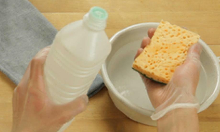 Hands holding a sponge and a bottle of vinegar above a bowl, preparing to apply vinegar to remove fabric glue from jeans.
